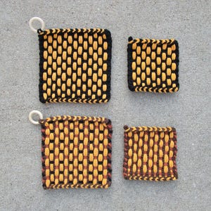 May include: Four square potholders in two sizes. Two feature a black and yellow woven pattern with a white ring attached. Two feature a brown and yellow woven pattern. The larger two potholders include a white ring for hanging on the top left corner.