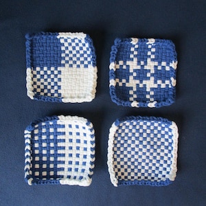 May include: Four square woven coasters in blue and white. Each coaster features a unique pattern, such as checkerboard and cross designs. The edges are finished with a blue or white border. The coasters are displayed on a dark blue surface.