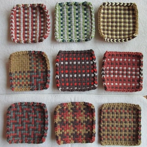 May include: Nine square woven pot holders in various colors, including red, green, brown, and gray. Each pot holder has a unique woven pattern and a braided edge. The pot holders are arranged on a white surface.