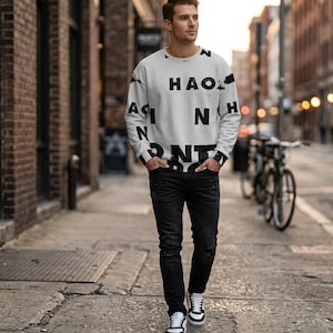 Può includere: Una persona indossa una felpa bianca con testo nero grande e jeans neri. La felpa ha le parole "HAO" e "N" stampate sopra. La persona indossa scarpe da ginnastica bianche e nere.