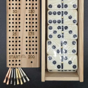 Puede incluir: Un tablero de cribbage de madera con agujeros para anotar y clavijas de metal, junto a una caja de madera que contiene dominós envueltos. El tablero de cribbage tiene números del 25 al 200. Los dominós son de color marfil con puntos negros.