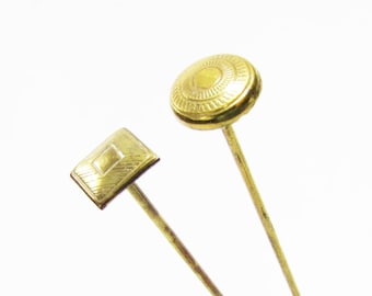 May include: Two gold-toned hat pins with decorative tops. One pin has a square top with a geometric design, and the other has a round top with a circular design.