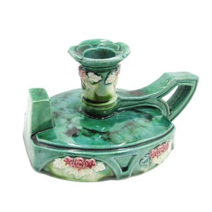May include: A glazed ceramic chamberstick candle holder in a mottled green finish. The piece features a central candle cup, a side handle, and raised relief floral accents in pink and white around the base and the neck of the candle holder.