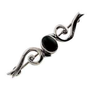 May include: A silver-toned brooch with an oval, dark-colored stone at its center. The brooch features a flowing, symmetrical design with swirling details and a pin closure. The overall shape is elongated and elegant.
