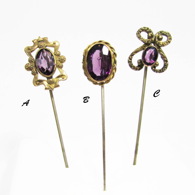 May include: Three vintage hat pins with purple glass stones. The pins are gold-toned and have different designs. The pins are labeled A, B, and C.
