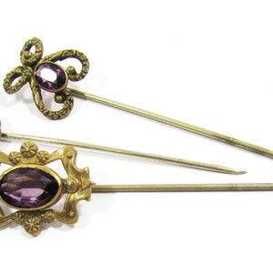 May include: Three antique hat pins with purple glass stones and gold-toned metal accents. The pins are arranged in a row on a white background.