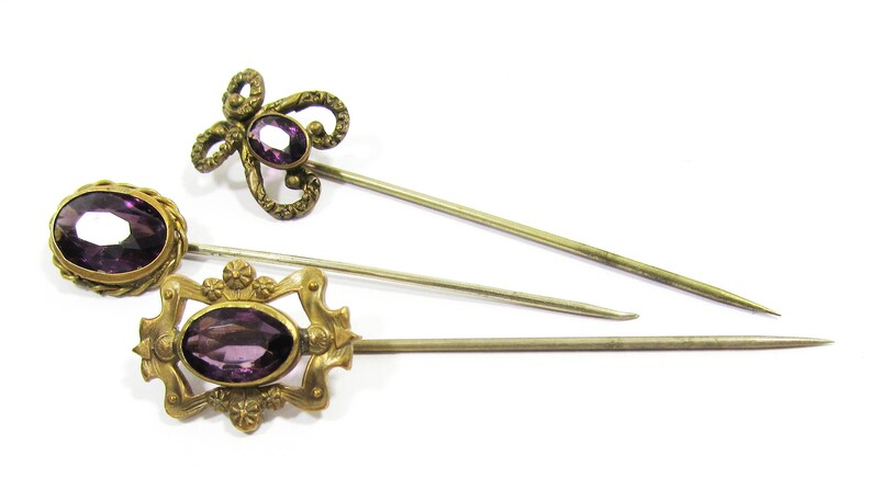 May include: Three antique hat pins with purple glass stones and gold-toned metal accents. The pins are arranged in a row on a white background.