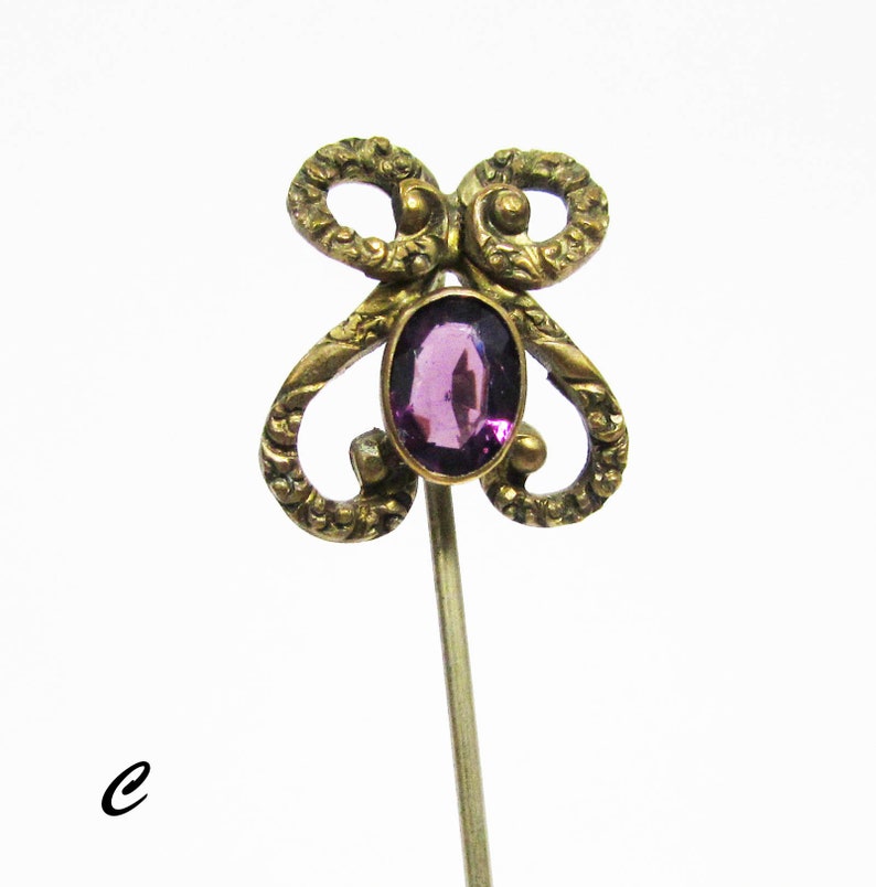 May include: A gold-toned pin with a purple gemstone in the center. The pin has a decorative design that resembles a butterfly.