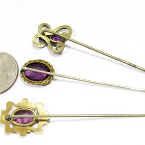 May include: Three antique hat pins with gold-toned metal settings and purple glass stones. The pins are arranged in a row on a white background.