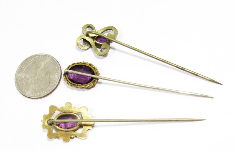 May include: Three antique hat pins with gold-toned metal settings and purple glass stones. The pins are arranged in a row on a white background.