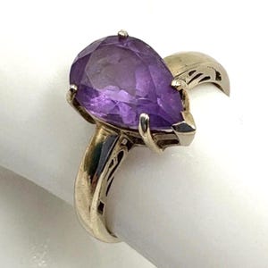 以下が含まれることがあります： 大きな洋梨型の紫色のファセットアメジスト宝石を特徴とするシルバーリング。 リングは、バンドと石を固定する爪に詳細な透かし彫りのデザインが施されています。 リングは白い表面に表示されています。