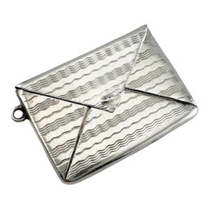 May include: A silver-toned metal charm shaped like a rectangular envelope. The surface features a repeating pattern of horizontal wavy lines etched across the front flap and body. A small circular jump ring is attached to the left side edge.