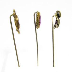 May include: Three gold-toned hair pins with purple glass stones. The pins have ornate designs and are on a white background.