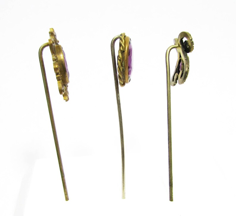 May include: Three gold-toned hair pins with purple glass stones. The pins have ornate designs and are on a white background.