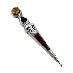 May include: A silver-toned kilt pin with a decorative design. The pin features a yellow gemstone at the top, a teal-colored stone, and a dark red inlay. The pointed end is detailed with a small ball.
