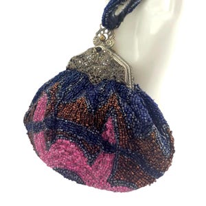 May include: A small, rounded beaded pouch featuring a geometric pattern in dark blue, copper, and bright pink glass beads. The bag has a silver-toned metal frame with intricate filigree detailing and a matching beaded handle at the top.