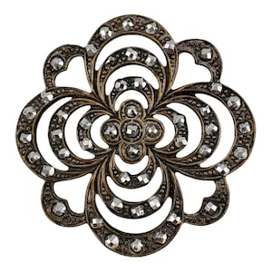 Peut inclure: Une broche circulaire présentant un design ajouré complexe avec une finition bronze vieilli. La structure métallique est ornée de nombreuses petites pierres facettées aux tons argentés, disposées selon un motif floral symétrique.