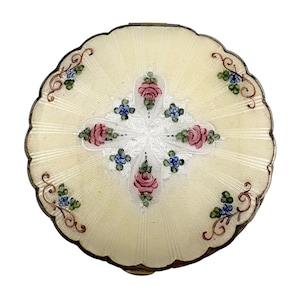 May include: A round, scalloped-edge compact case featuring a cream-colored guilloché enamel surface with a radiating sunburst pattern. The center displays a white cross-shaped floral motif with pink roses and small blue flowers, accented by gold.