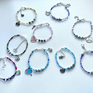 May include: A selection of beaded bracelets in shades of yellow, blue, red, and purple. Each bracelet has a unique charm, like a musical note, a heart, or a cartoon character. The bracelets are presented on a white surface.