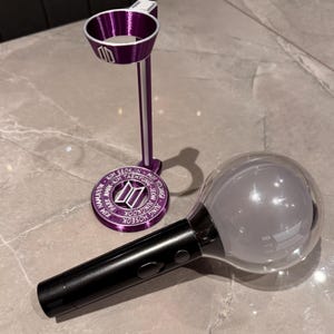 May include: A purple light stick stand and a black and clear light stick. The stand has a circular base with engraved names and a logo. The light stick has a clear orb and the letters "BTS".