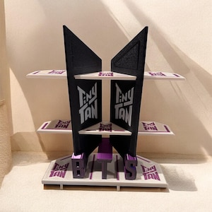 May include: A black and purple shelf unit with three levels. The design features the text "TINY TAN" and "BTS". The shelf is made of a robust material and is suitable for displaying small items. Measurements are in centimeters.