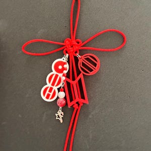 May include: A red and white charm with a silver clasp. The charm features a red knot, a red and white circular design, a red logo, a star charm, and red beads. The charm is attached to a silver clasp.