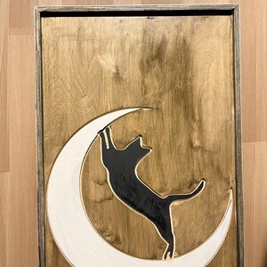 May include: Wooden wall art featuring a black cat climbing a white crescent moon against a stained wood background. The artwork is framed in a thin, dark-toned wooden frame. The cat is reaching upwards.