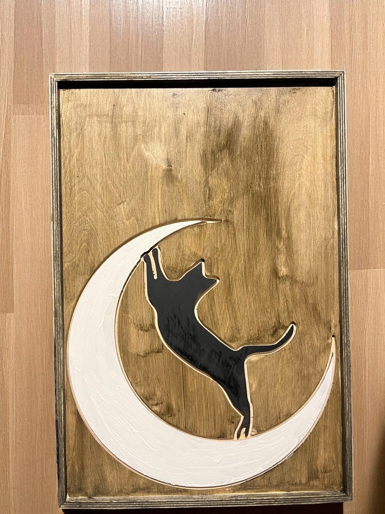 May include: Wooden wall art featuring a black cat climbing a white crescent moon against a stained wood background. The artwork is framed in a thin, dark-toned wooden frame. The cat is reaching upwards.
