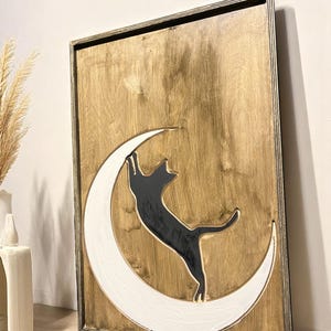 May include: Wooden wall art featuring a black cat silhouette reaching for a white crescent moon. The cat and moon are set against a natural wood grain background within a framed border. A decorative piece for cat lovers.
