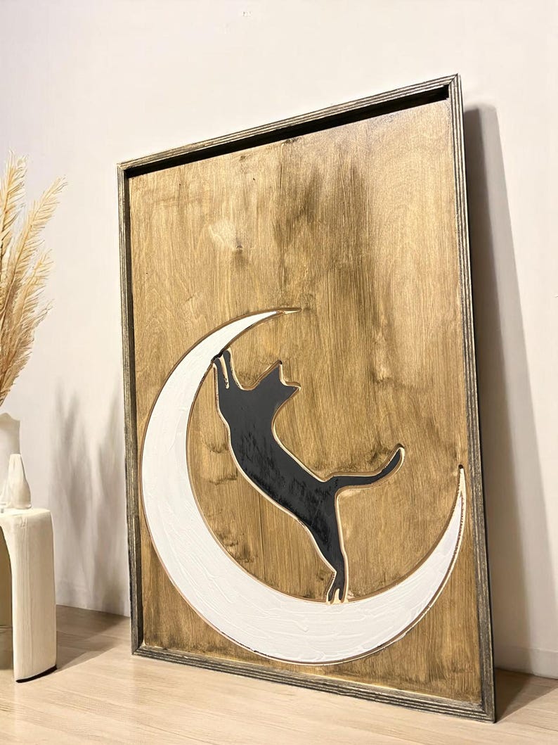 May include: Wooden wall art featuring a black cat silhouette reaching for a white crescent moon. The cat and moon are set against a natural wood grain background within a framed border. A decorative piece for cat lovers.