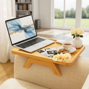 May include: A rectangular bamboo sofa arm tray table with a laptop, a white ceramic teacup on a saucer with two cookies, and a small white vase containing three pale yellow roses. The laptop screen displays an abstract blue and gold design.