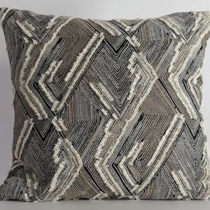 May include: Square decorative pillow with a geometric pattern in shades of black, gray, and beige. The design features a series of angled lines and diamond shapes, with textured, fringed accents. The pillow is a neutral home decor item.