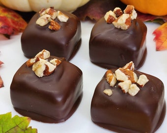 May include: Four square chocolate candies topped with chopped pecans. The candies are arranged on a white plate with fall leaves around the edges.