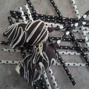 May include: Two chocolate-covered treats with white drizzle, resting on a bed of black and white polka dot straws. The treats have a rectangular shape and are decorated with white stripes. The straws are a mix of black with white polka dots and white with black polka dots.