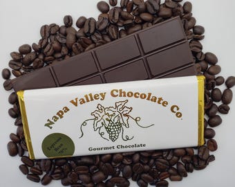 May include: A white chocolate bar with gold foil wrapping, featuring the text "Napa Valley Chocolate Co." and "Gourmet Chocolate". The bar is surrounded by coffee beans and has a label that says "Espresso Bean 70%".