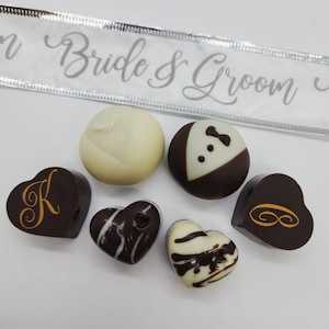 May include: Assortment of six chocolate candies in heart and round shapes. The chocolates are decorated with white, brown, and gold designs. The chocolates are arranged on a white surface. The text "Bride & Groom" is visible on a white ribbon.