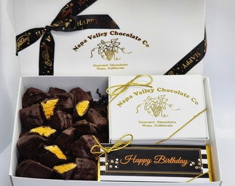 May include: A white gift box with a black ribbon tied in a bow. The box contains a selection of chocolate honeycomb candy and a small white box with a gold ribbon. The small box has a gold logo that says "Napa Valley Chocolate Co." and the text "Gourmet Chocolates Napa, California". The small box also has a black and white gift tag that says "Happy Birthday".