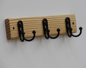 May include: A wooden wall-mounted coat rack with three black metal hooks. The rack is made of light-colored wood with visible grain and is mounted on a white wall. The hooks have a rounded design.