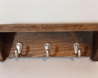 May include: A rustic wooden shelf with three metal hooks. The shelf is made of dark stained wood and has a simple, functional design. The hooks are silver-colored and attached with screws. Perfect for entryway or mudroom.