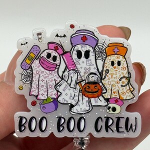 May include: A glittery acrylic badge reel featuring three ghost characters dressed as medical professionals with nurse caps, a pink syringe, a bandage, and a pumpkin bucket. The design includes medical icons and the text BOO BOO CREW in black.
