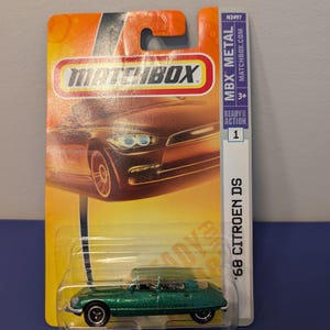 May include: A green die-cast metal '68 Citroen DS toy car on a Matchbox card. The car is a metallic green colour with black wheels. The card is orange and yellow with the Matchbox logo and the text "MBX METAL".