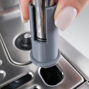 May include: A gray, cylindrical 3D-printed part being inserted into a stainless steel appliance. The part has a circular base and a slot to accommodate a metal tube. The background is a close-up of the appliance's surface.