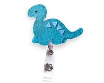 May include: A teal blue felt dinosaur with white felt accents is attached to a white retractable badge holder.