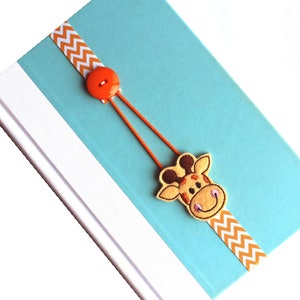 May include: A blue book with a white border and a yellow and white chevron ribbon bookmark with a giraffe face and an orange button.