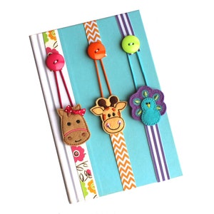 May include: Three felt animal bookmarks with buttons and ribbon. The bookmarks feature a horse, giraffe, and peacock. The horse is brown with a pink bow, the giraffe is orange and brown, and the peacock is purple and blue.