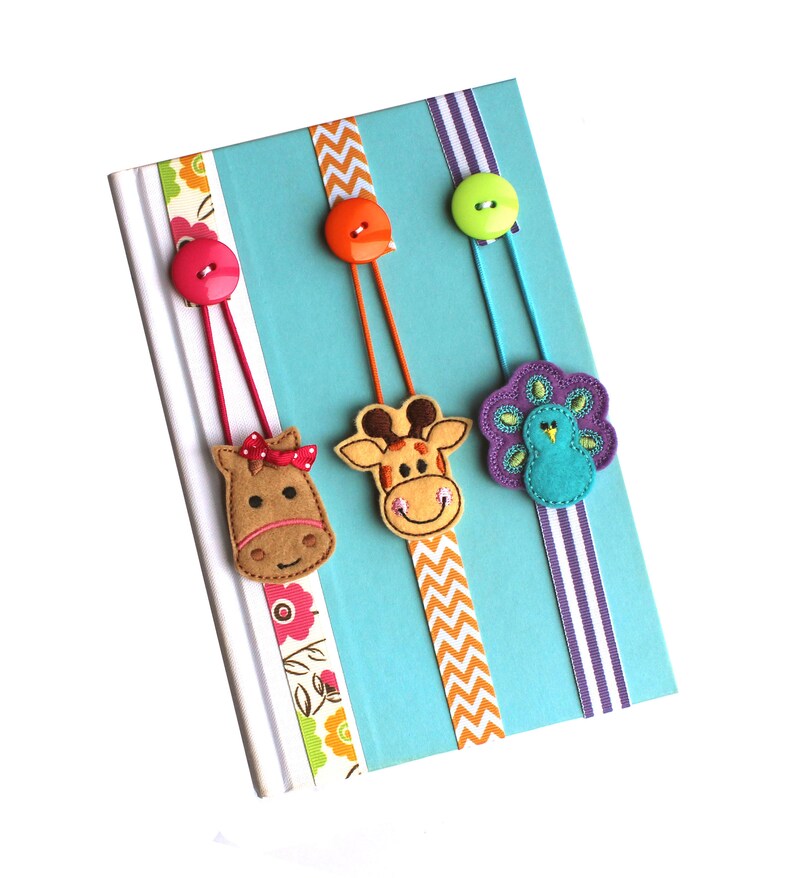 May include: Three felt animal bookmarks with buttons and ribbon. The bookmarks feature a horse, giraffe, and peacock. The horse is brown with a pink bow, the giraffe is orange and brown, and the peacock is purple and blue.