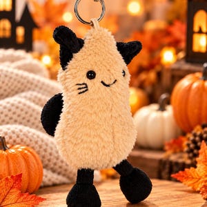 May include: A plush keychain shaped like a pear-like creature with tan fuzzy fur, black corduroy ears, black corduroy feet, and black embroidered facial features including eyes, a small smile, and whiskers, attached to a silver metal keyring.