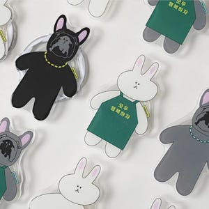 May include: A selection of cartoon phone grips. The designs include a black bulldog with a gold necklace, a white bunny, and a grey bulldog. The bunny grips wear a green apron with Korean text.