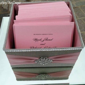Puede incluir: Una caja de dos niveles con un exterior de tela gris y rosa. La caja está llena de programas de ceremonia de boda de color rosa. El nivel superior tiene un adorno de pedrería y una cinta rosa. Los programas tienen el texto "The Wedding Ceremony Uniting Nicole Strand and Richard Lindquist".