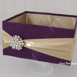 May include: A square box with a purple fabric exterior and a gold satin ribbon. The box is decorated with a large pearl brooch.